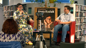 Zdjęcie przedstawia rozmawiającą autorkę, Annę Kasiuk z prowadzącym spotkanie bibliotekarzem na małej scenie wewnątrz biblioteki